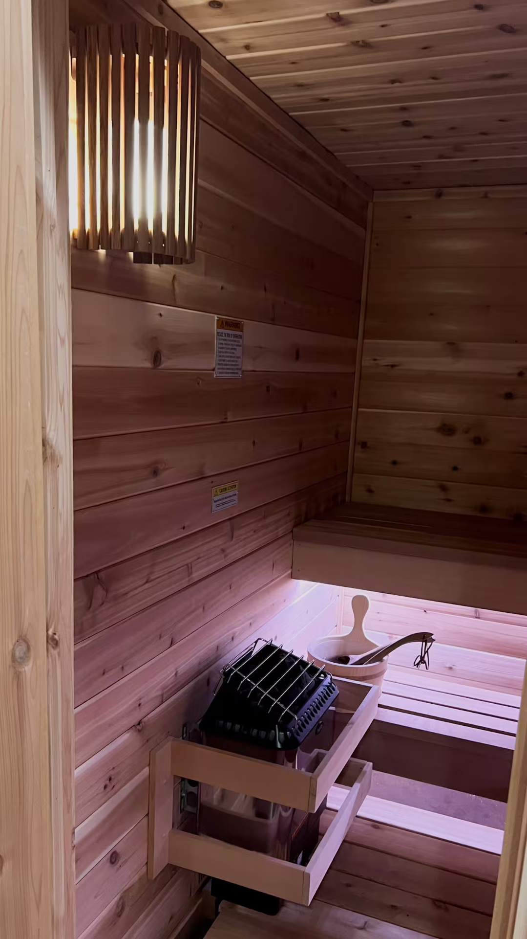 Inside the cedar sauna with heater, bucket, and warm wood lighting