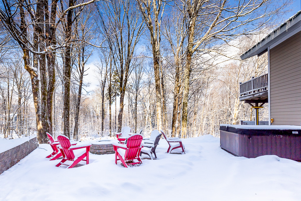 Outdoor fire pit with red Adirondack chairs and hot tub beside snowy birch woods
